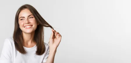 13 Ways to Make Your Hair Stronger and Healthier - portrait flirty beautiful girl playing with hair smiling camera coquettish standing white scaled